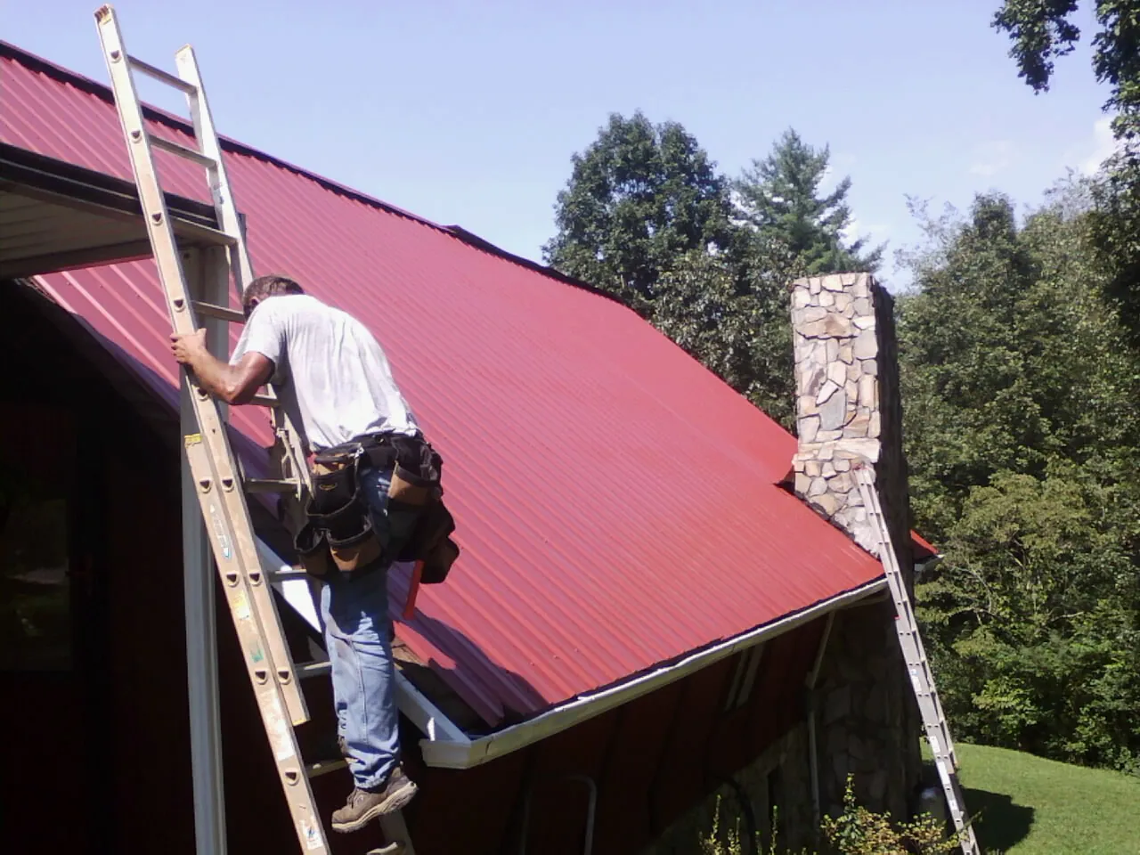 Roofer on a ladder beside a red metal roof and stone chimney — Soffit & Fascia Repair by Roofing Brothers in Gloversville